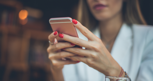 A woman in a white blazer using a smartphone with red nails in a cozy setting.